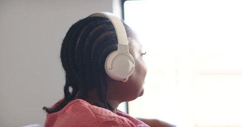 African american woman wearing white headphones listening by sunlit window, pink top, cornrows