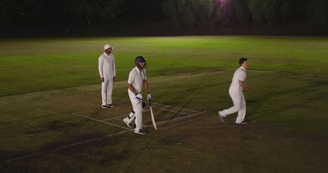Night cricket match under floodlights with diverse players batting, running on grass pitch