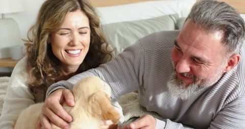 Smiling mature couple petting cream puppy on cozy bed wearing knit sweaters