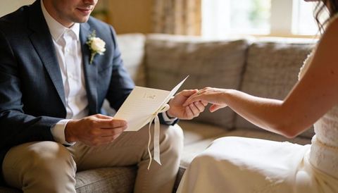 Groom reading vows while holding bride's ringed hand on cozy sofa, intimate wedding moment