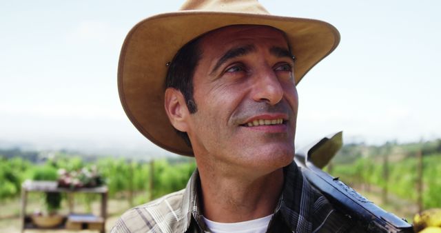 Middle-aged farmer smiling while standing in vineyard, wearing a hat, on sunny day. Background showcases grapevines and agricultural activity. Perfect for promoting farming, agriculture, rural lifestyle, sustainable farming practices, vineyard tours, and farmer profiles or stories.