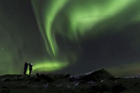 Couple watching vibrant green aurora borealis dancing over snowy rocky arctic landscape