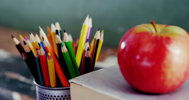 A vibrant collection of colored pencils in a holder on a wooden desk next to a red apple and a book. This image can be used for back-to-school promotions, education materials, classroom decor ideas, or stationery advertisements.