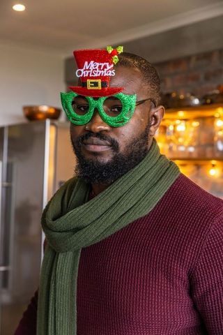 African american man wearing merry christmas glitter glasses and scarf in cozy kitchen
