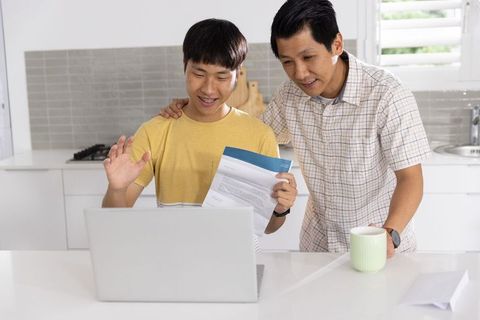 Asian father and teenage son reviewing documents during video call on laptop in kitchen