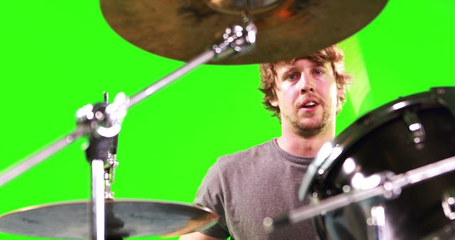 A young Caucasian male musician plays the drums in a studio with a green screen background, with copy space. His focused expression and dynamic posture capture the energy and intensity of a drumming performance.