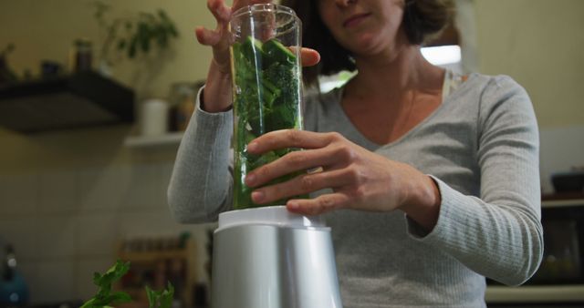 Woman blending fresh green ingredients in a modern kitchen, emphasizing healthy living and nutritious meals. Ideal for articles on wellness, nutrition, healthy cooking, vegetarian or vegan recipes. Perfect for use in advertisements for kitchen appliances, health food brands, or culinary blogs.