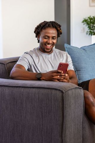 African man relaxing on gray sofa smiling while using red smartphone for messages and entertainment