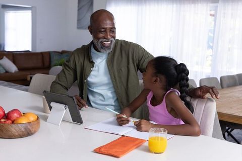 Grandfather tutoring granddaughter at kitchen counter using tablet and notebook