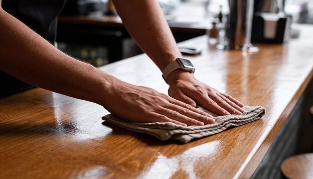 Bartender cleaning shiny wooden bar counter with cloth and smartwatch