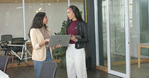 Diverse female coworkers collaborating with laptop and tablet in modern glass office space
