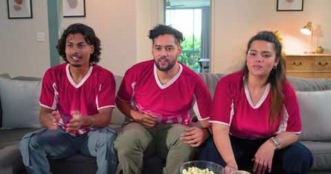 Diverse friends cheering during game night on sofa wearing red jerseys with popcorn bowl