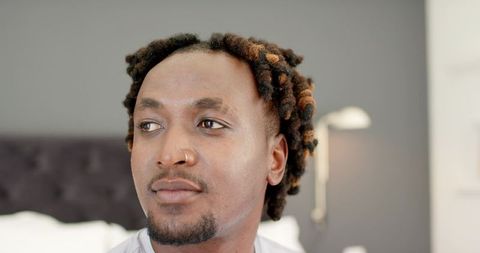 African man sitting in bedroom looking thoughtful wearing light shirt and nose ring with locs