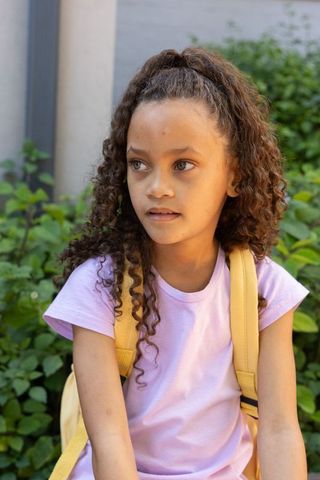 Pensive schoolgirl sitting outdoors with yellow backpack straps, curly hair, purple shirt