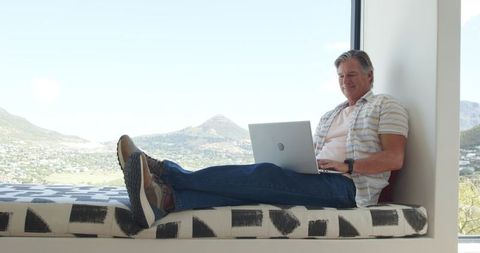 Mature man working remotely on laptop from sunlit window bench with panoramic valley view