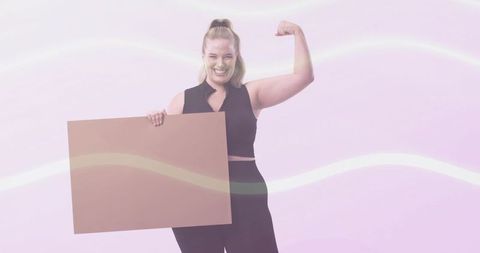 Confident woman holding blank cardboard sign flexing arm and smiling on pink backdrop