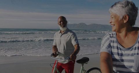 Senior couple cycling and chatting along sunny beach shoreline, smiling and enjoying sea breeze