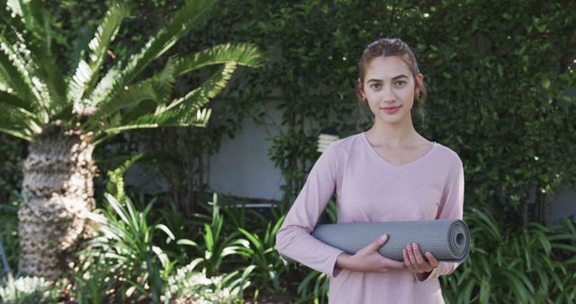 Young woman holding a rolled yoga mat in an outdoor garden with lush greenery. Ideal for content related to fitness, healthy lifestyle, yoga practice, outdoor activities, and mindfulness.