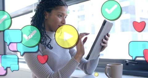 Young woman tapping tablet by window with floating social media icons and coffee mug