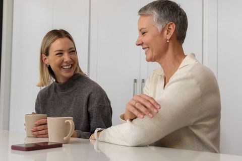 Mature mother and adult daughter sharing coffee and laughing in modern kitchen