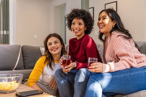 Multicultural friends laughing and sharing drinks on cozy living room sofa
