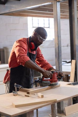 Carpenter sanding wood with red orbital sander on workbench in bright workshop