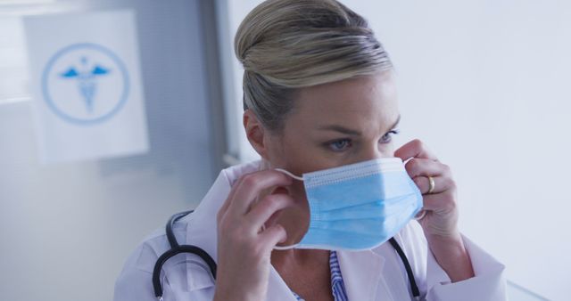 Female doctor is adjusting a blue face mask in a hospital corridor. She is wearing a white medical coat and has a stethoscope around her neck. This can be used for health-related contexts, focusing on safety, hospital routines, doctor professionalism, and COVID-19 precautions.