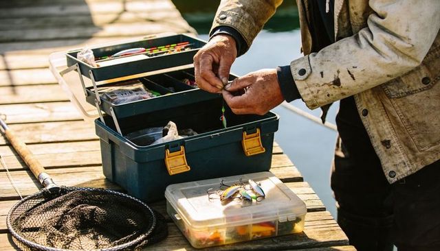 Angler preparing fishing lures on sunlit wooden dock tackle box net close-up