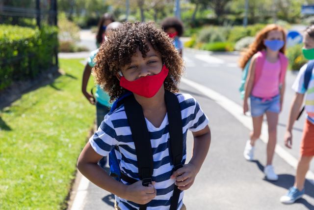 African American boy wearing a red face mask and carrying a backpack walking on a footpath outdoors. Other children can be seen in the background, also wearing backpacks, practicing social distancing. Useful for illustrating themes of school reopening during the coronavirus pandemic, health and safety measures, education in the new normal, and children's activities during COVID-19.