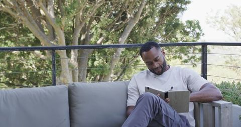 Mature man reading and writing on sunlit balcony sofa with notebook and pen, relaxed patio