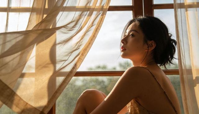 Contemplative asian woman sitting on wooden windowsill at golden hour with flowing curtains