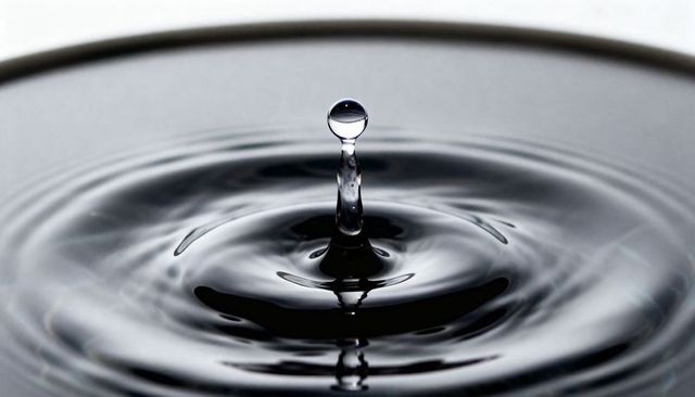 Rising water droplet balancing on liquid column with concentric ripples and glossy reflection