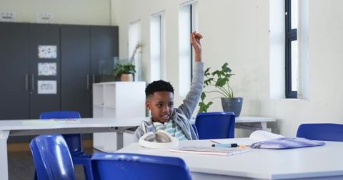 Young student raising hand at classroom table with headphones, books and pencil case