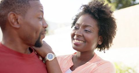 African american couple smiling and chatting in sunlit garden, woman touching man's watch