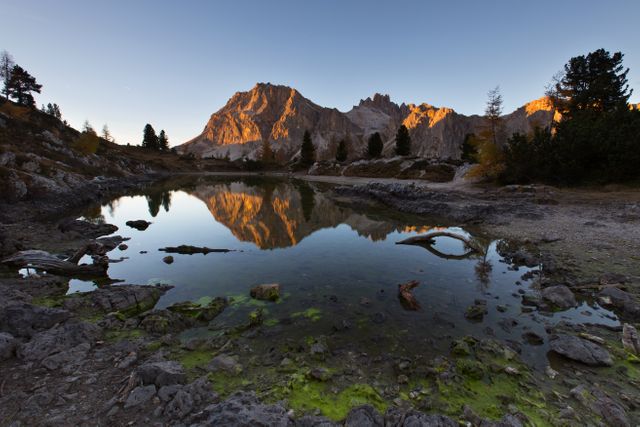 Scenic image of a sunset casting a warm glow over mountains reflected in a calm Alpine lake. Ideal for use in travel brochures, nature-themed blogs, outdoor adventure advertisements, and decor for promoting relaxation and tranquility. The serene water and natural landscape can inspire a sense of peace and escapism.