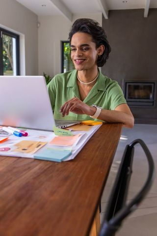 Asian man working on laptop at wooden table with sticky notes, green shirt, smartwatch