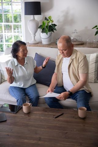 Senior asian couple discussing bills and financial documents on sofa with tablets, coffee