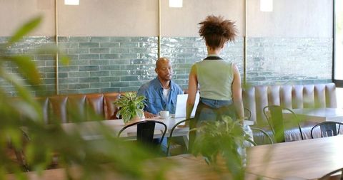 Cafe meeting with laptop and green plants, man seated, woman standing at banquette