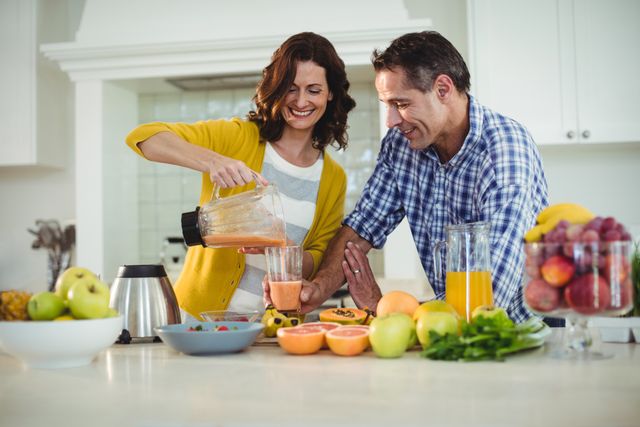 Happy couple preparing a smoothie in a bright kitchen. They are surrounded by fresh fruits and vegetables, indicating a healthy lifestyle. This image can be used for promoting healthy eating, family bonding, morning routines, and domestic life.