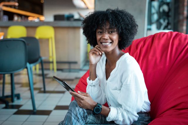African American woman sitting on a red beanbag in a modern cafe, using a smartphone. She is smiling and appears relaxed. This image is ideal for promoting digital nomad lifestyles, modern technology use, coworking spaces, and urban leisure activities.