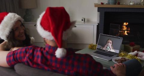Couple Wearing Santa Hats on Video Call by Fireplace with Laptop and Holiday Cookies