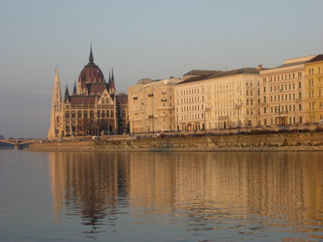 Hungarian Parliament Building and adjacent historic structures as seen from across the Danube River during golden hour. Illuminated by the setting sun, building reflections create a serene scene on the calm water. Ideal for use in travel blogs, architecture portfolios, tourism promotions, and European culture presentations.