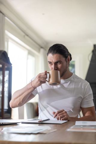 Man in 30s sipping coffee while reviewing documents at sunlit home workspace