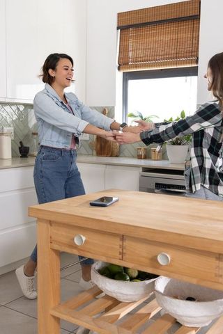 Young women holding hands and chatting at wooden kitchen island, cozy daylight moment