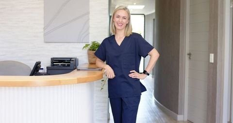A confident nurse in blue scrubs is seen using a tablet at a reception desk in a modern medical clinic. The setting exudes professionalism with its clean and organized appearance. Ideal for use in healthcare marketing materials, medical brochures, or any context highlighting medical technology integration and patient-care facilities.