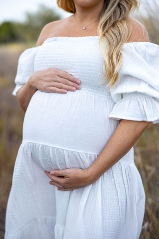 Pregnant woman in white off-shoulder dress embracing belly outdoors