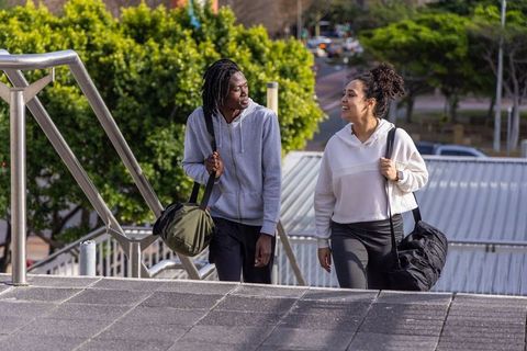 Diverse friends walking up urban stairs carrying gym duffels chatting on sunny day