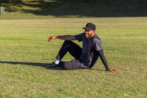 Athletic man relaxing on sunlit park grass wearing cap and trainers, smiling and leaning back