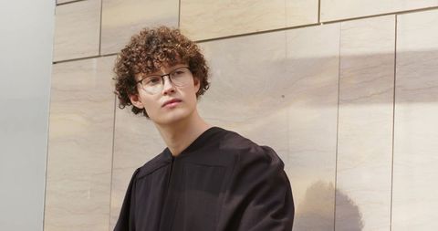 Contemplative graduate wearing black academic robe and round glasses against modern stone wall