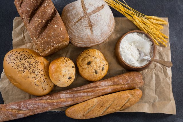 Assorted bread loaves including baguettes, sourdough, and whole grain bread arranged on a rustic background with wheat grains and a bowl of flour. Ideal for use in bakery promotions, food blogs, recipe websites, and healthy eating campaigns.
